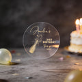 A round clear sticker with gold foiling saying 'Amelia's 21st Birthday Thank you for coming' and a champagne bottle illustration, with blurred birthday balloons and a lit cake in the background.
