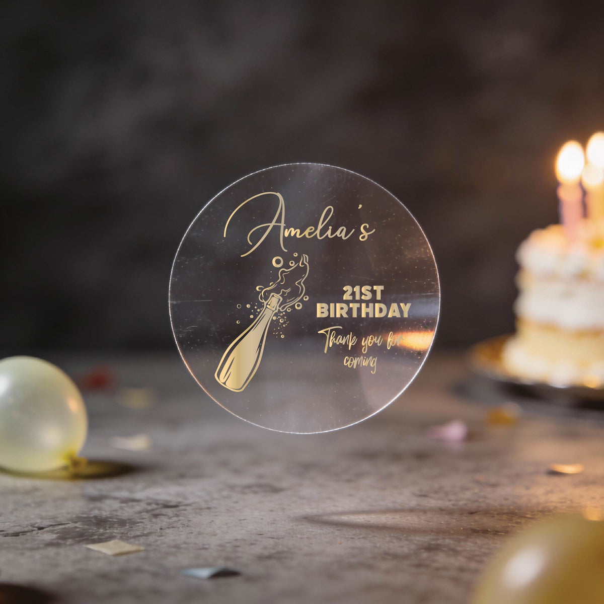 A round clear sticker with gold foiling saying 'Amelia's 21st Birthday Thank you for coming' and a champagne bottle illustration, with blurred birthday balloons and a lit cake in the background.