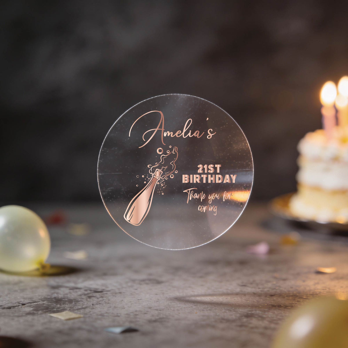A round clear sticker with rose gold foiling saying 'Amelia's 21st Birthday Thank you for coming' and a champagne bottle illustration, with blurred birthday balloons and a lit cake in the background.