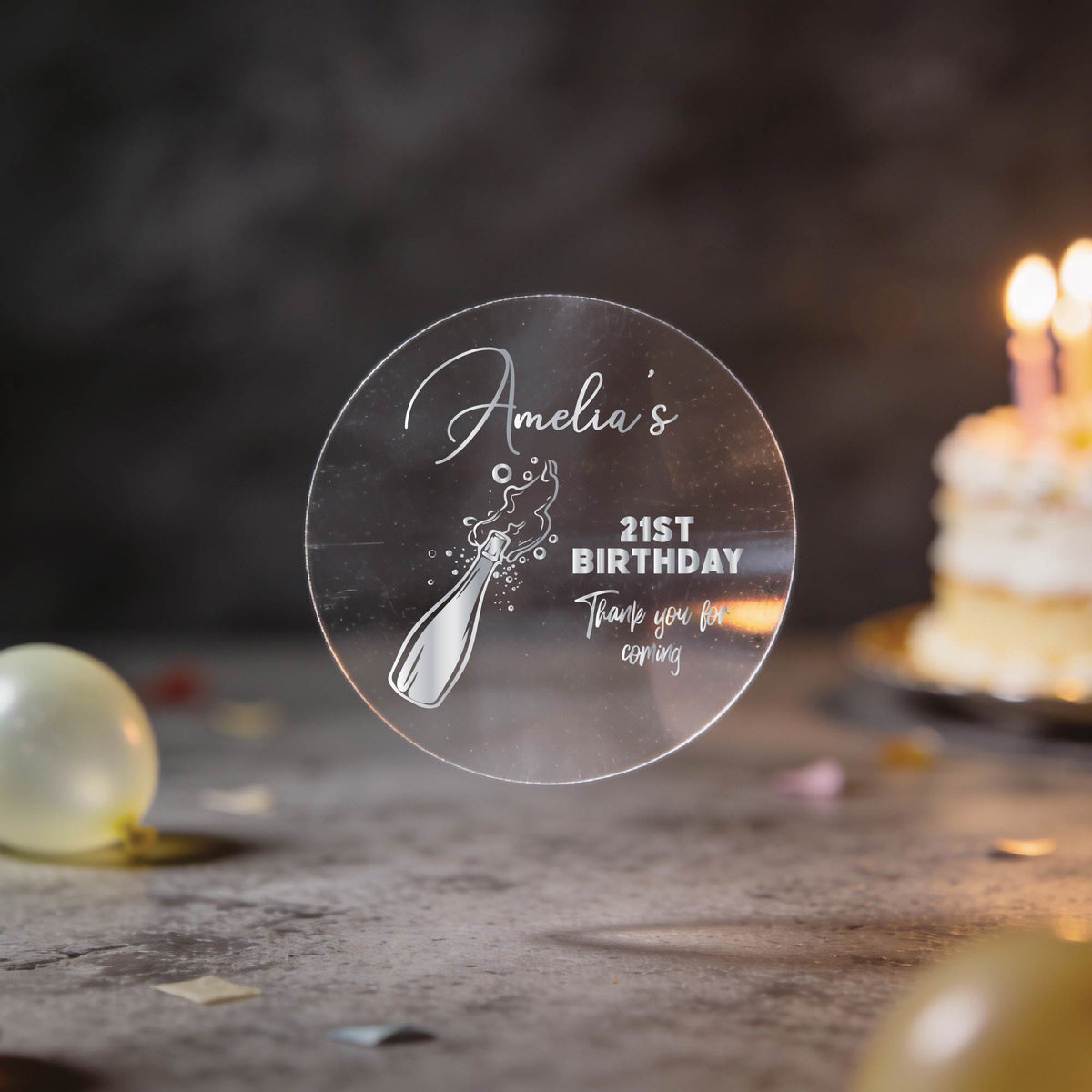A round clear sticker with silver foiling saying 'Amelia's 21st Birthday Thank you for coming' and a champagne bottle illustration, with blurred birthday balloons and a lit cake in the background.