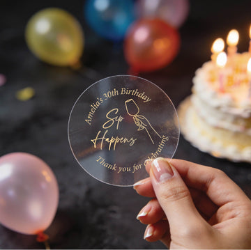 A clear round gold foil sticker with an image showing a hand holding a wine glass and the text 'Sip Happens', 'Amanda's 30th Birthday' & 'Thank you for celebrating'. The background is a dark birthday scene with blurred balloons and a lit cake.