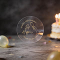 A clear round gold foil sticker with an image showing a hand holding a wine glass and the text 'Sip Happens', 'Amanda's 30th Birthday' & 'Thank you for celebrating'. The background is a dark birthday scene with blurred balloons and a lit cake.