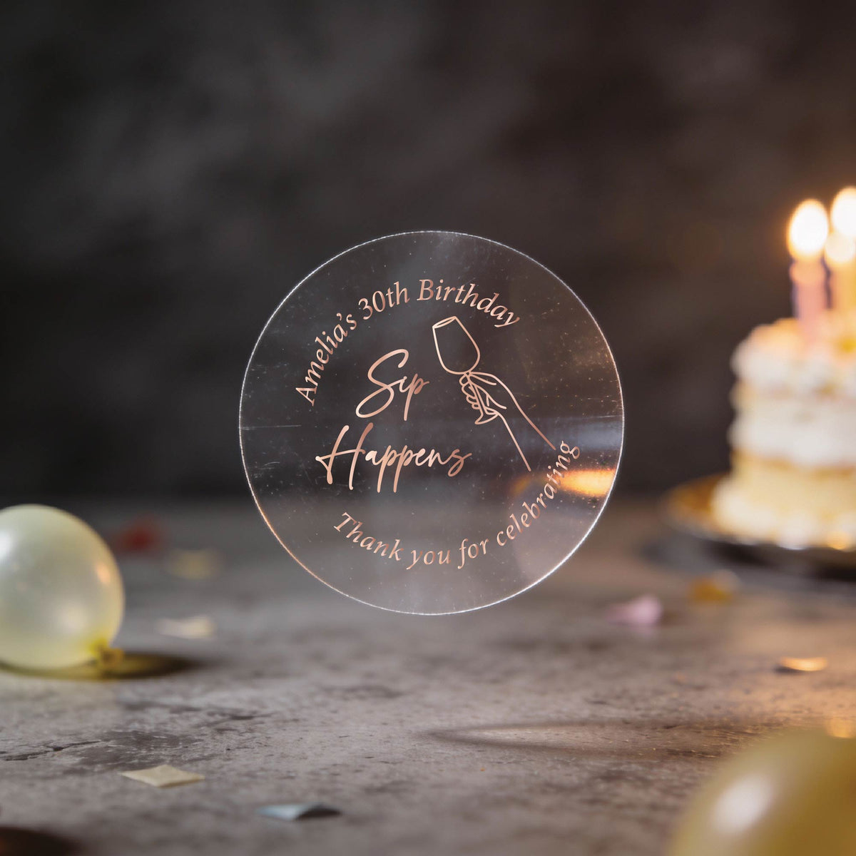 A clear round rose gold foil sticker with an image showing a hand holding a wine glass and the text 'Sip Happens', 'Amanda's 30th Birthday' & 'Thank you for celebrating'. The background is a dark birthday scene with blurred balloons and a lit cake.