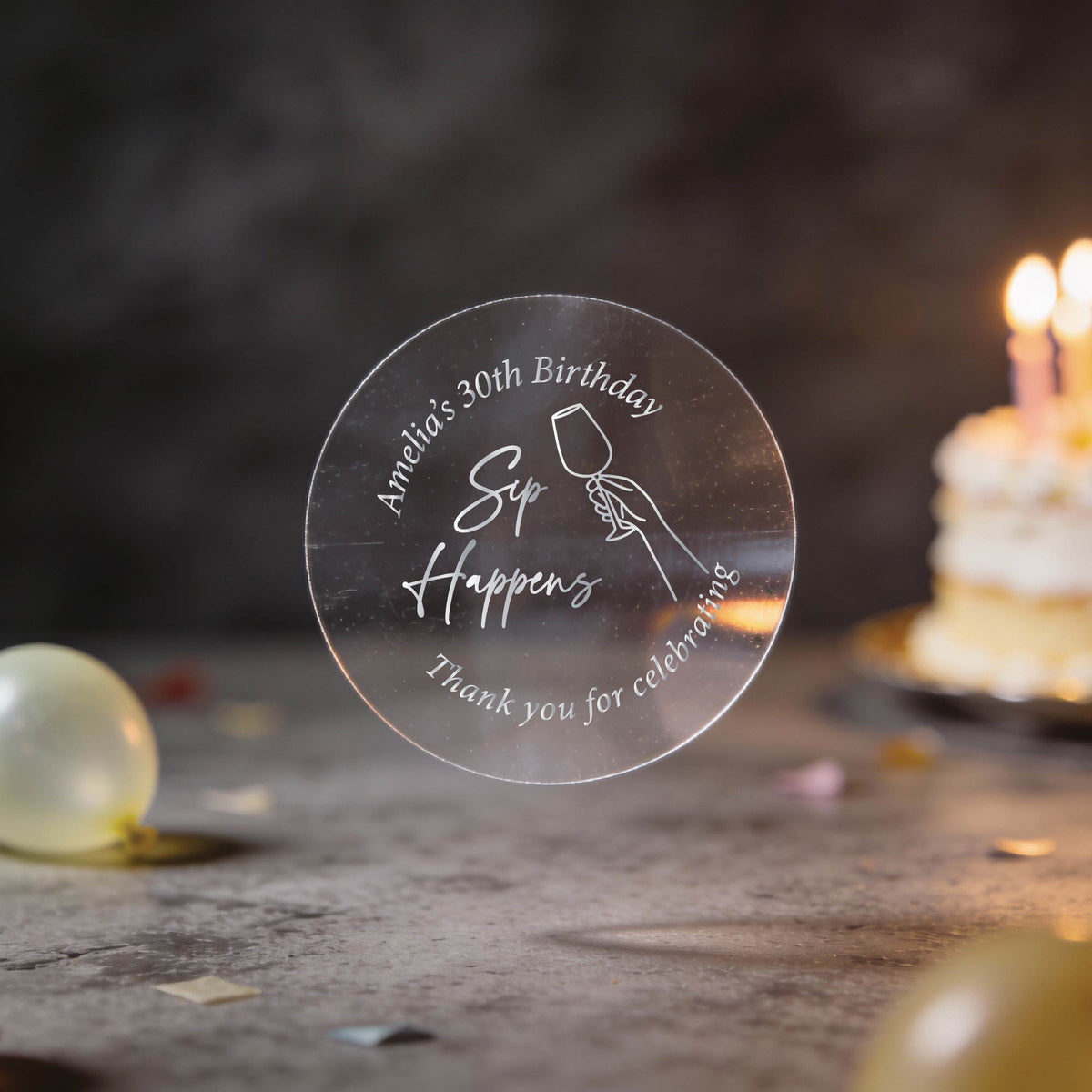 A clear round silver foil sticker with an image showing a hand holding a wine glass and the text 'Sip Happens', 'Amanda's 30th Birthday' & 'Thank you for celebrating'. The background is a dark birthday scene with blurred balloons and a lit cake.
