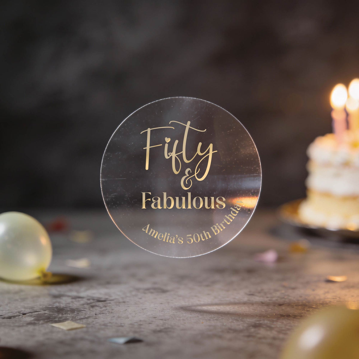 A clear round with gold foil text reading 'Fifty & Fabulous' & 'Amelia's 50th Birthday'. 
