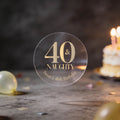 A clear round sticker with gold foil lettering that reads '40 & NAUGHTY Sarah's 40th Birthday'. The background is a dark, blurred birthday scene with balloons and a lit cake.