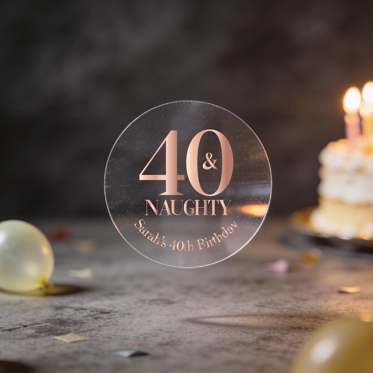 A clear round sticker with rose gold foil lettering that reads '40 & NAUGHTY Sarah's 40th Birthday'. The background is a dark, blurred birthday scene with balloons and a lit cake.