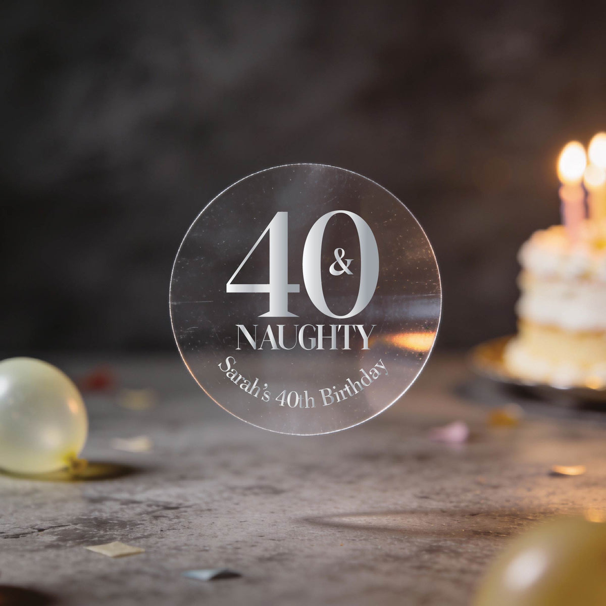 A clear round sticker with silver foil lettering that reads '40 & NAUGHTY Sarah's 40th Birthday'. The background is a dark, blurred birthday scene with balloons and a lit cake.