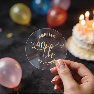 A hand holds a clear round sticker with gold foil text reading 'AMELIA'S 40th Birthday" and the date underneath. A lovely rose bud design in the middle to make these stickers extra special.