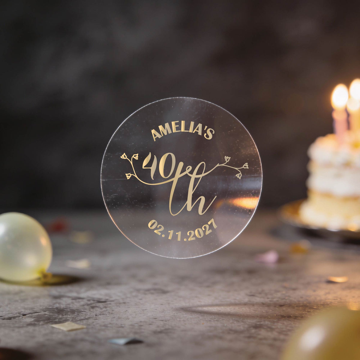 A hand holds a clear round sticker with gold foil text reading 'AMELIA'S 40th Birthday" and the date underneath. A lovely rose bud design in the middle to make these stickers extra special.
