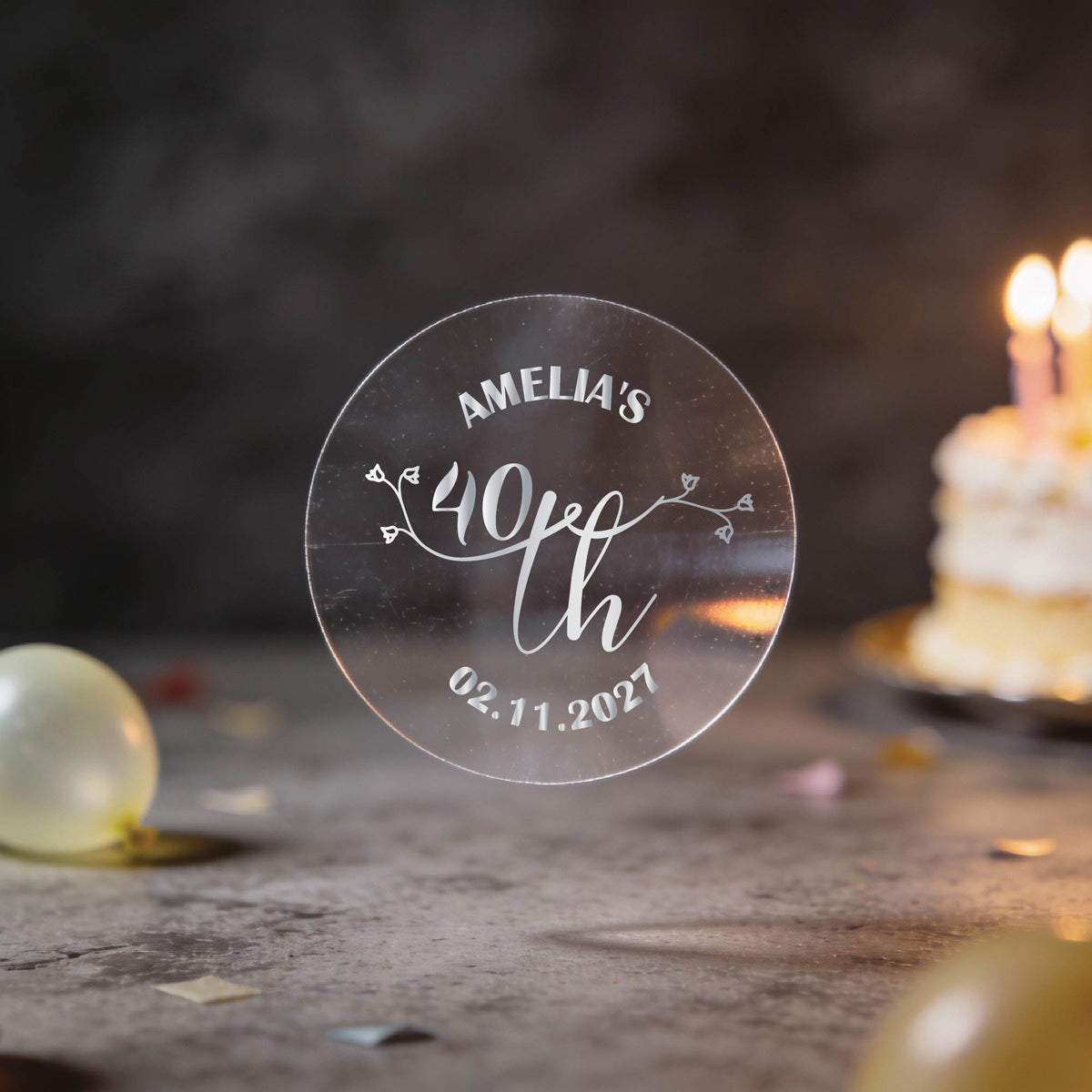 A hand holds a clear round sticker with silver foil text reading 'AMELIA'S 40th Birthday" and the date underneath. A lovely rose bud design in the middle to make these stickers extra special.