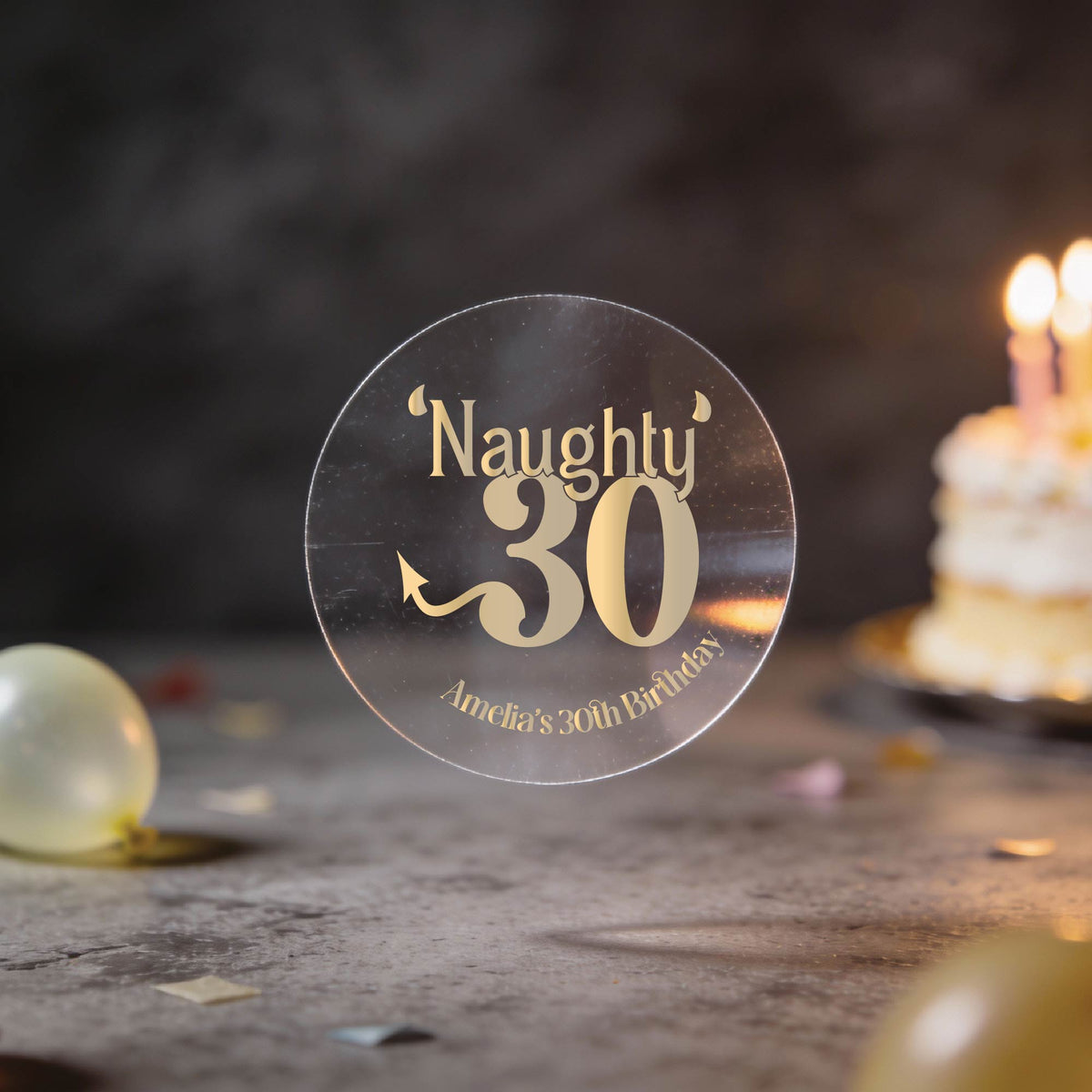 A clear round sticker with gold foil lettering that reads "Naughty 30" and "Amelia's 30th Birthday" against a dark party background with balloons and a birthday cake.