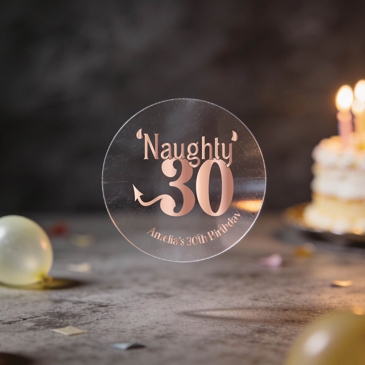 A clear round sticker with rose gold foil lettering that reads "Naughty 30" and "Amelia's 30th Birthday" against a dark party background with balloons and a birthday cake.