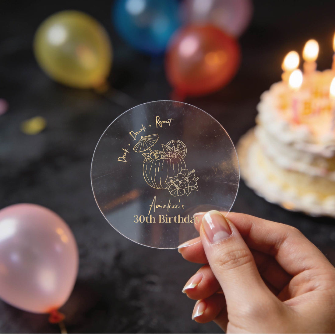 A hand holds a clear round sticker with gold foil text reading 'AMELIA'S 30th Birthday and 'Drink, Drunk & Repeat. A coconut cocktail drink design in the middle.