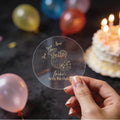 A hand holds a clear round sticker with gold foil text reading 'AMELIA'S 30th Birthday and 'Drink, Drunk & Repeat. A coconut cocktail drink design in the middle.