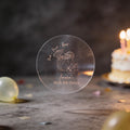 A hand holds a clear round sticker with rose gold foil text reading 'AMELIA'S 30th Birthday and 'Drink, Drunk & Repeat. A coconut cocktail drink design in the middle.