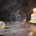 A hand holds a clear round sticker with silver gold foil text reading 'AMELIA'S 30th Birthday and 'Drink, Drunk & Repeat. A coconut cocktail drink design in the middle.