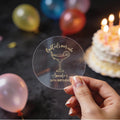 A hand holds a clear gold foil round sticker with a cocktail glass and the text 'Cocktails over cake' and 'Amanda's 30th Birthday'. The background is a dark birthday scene with blurred balloons and a lit cake.