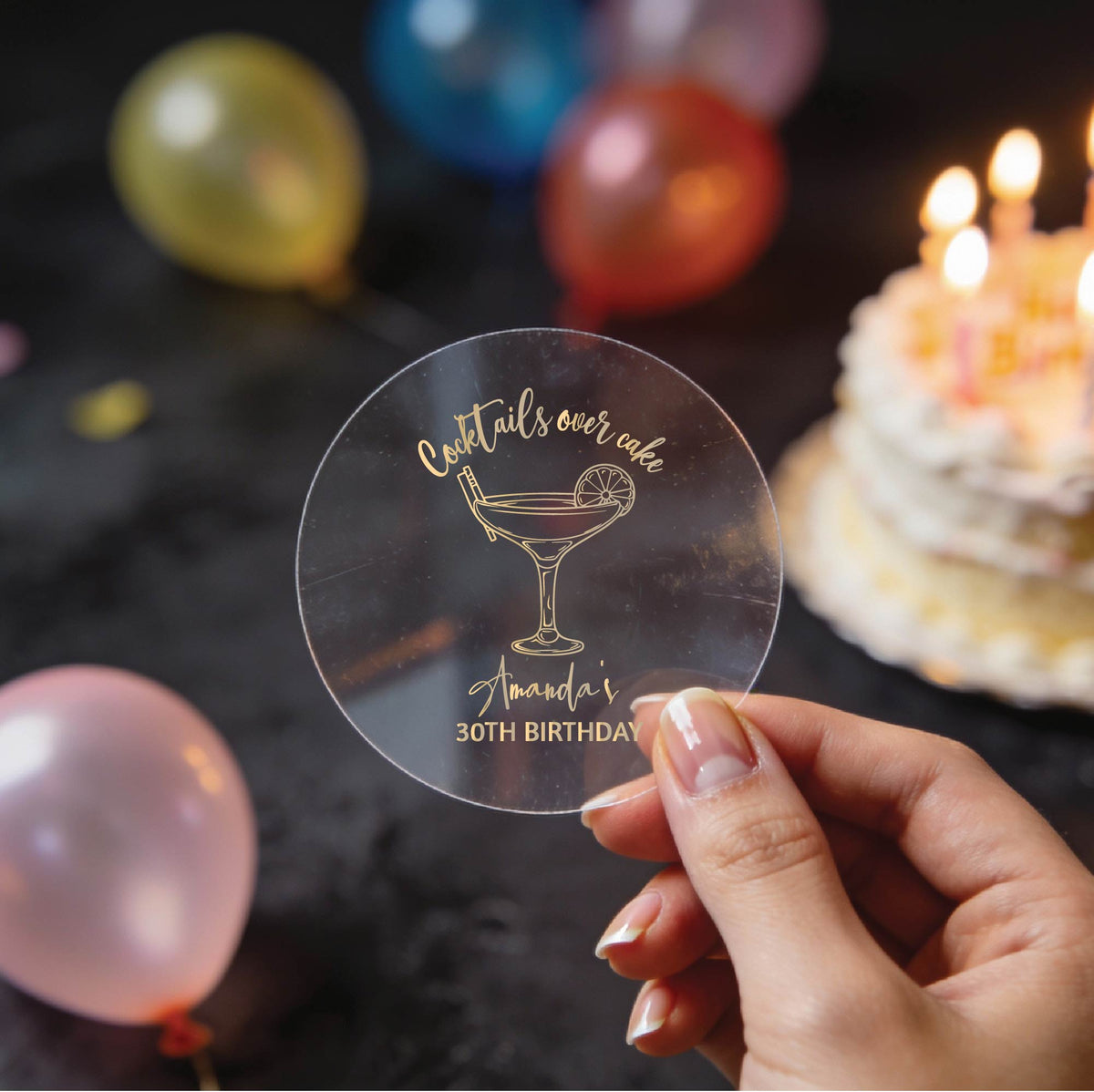 A hand holds a clear gold foil round sticker with a cocktail glass and the text 'Cocktails over cake' and 'Amanda's 30th Birthday'. The background is a dark birthday scene with blurred balloons and a lit cake.