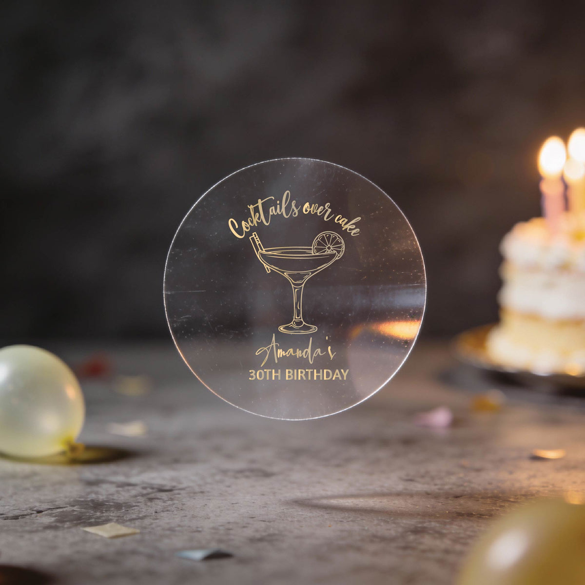 A clear gold foil round sticker with a cocktail glass and the text 'Cocktails over cake' and 'Amanda's 30th Birthday'. The background is a dark birthday scene with blurred balloons and a lit cake.