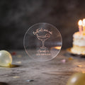 A clear rose gold foil round sticker with a cocktail glass and the text 'Cocktails over cake' and 'Amanda's 30th Birthday'. The background is a dark birthday scene with blurred balloons and a lit cake.