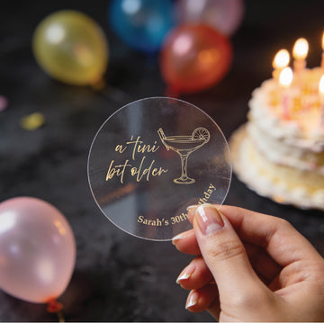 A hand holding a round clear sticker with the words in gold foil saying 'a tini bit older' and a martini glass illustration, with 'Sarah's 30th birthday' printed at the bottom. The background is a dark party setting with blurred balloons and a birthday cake with lit candles.