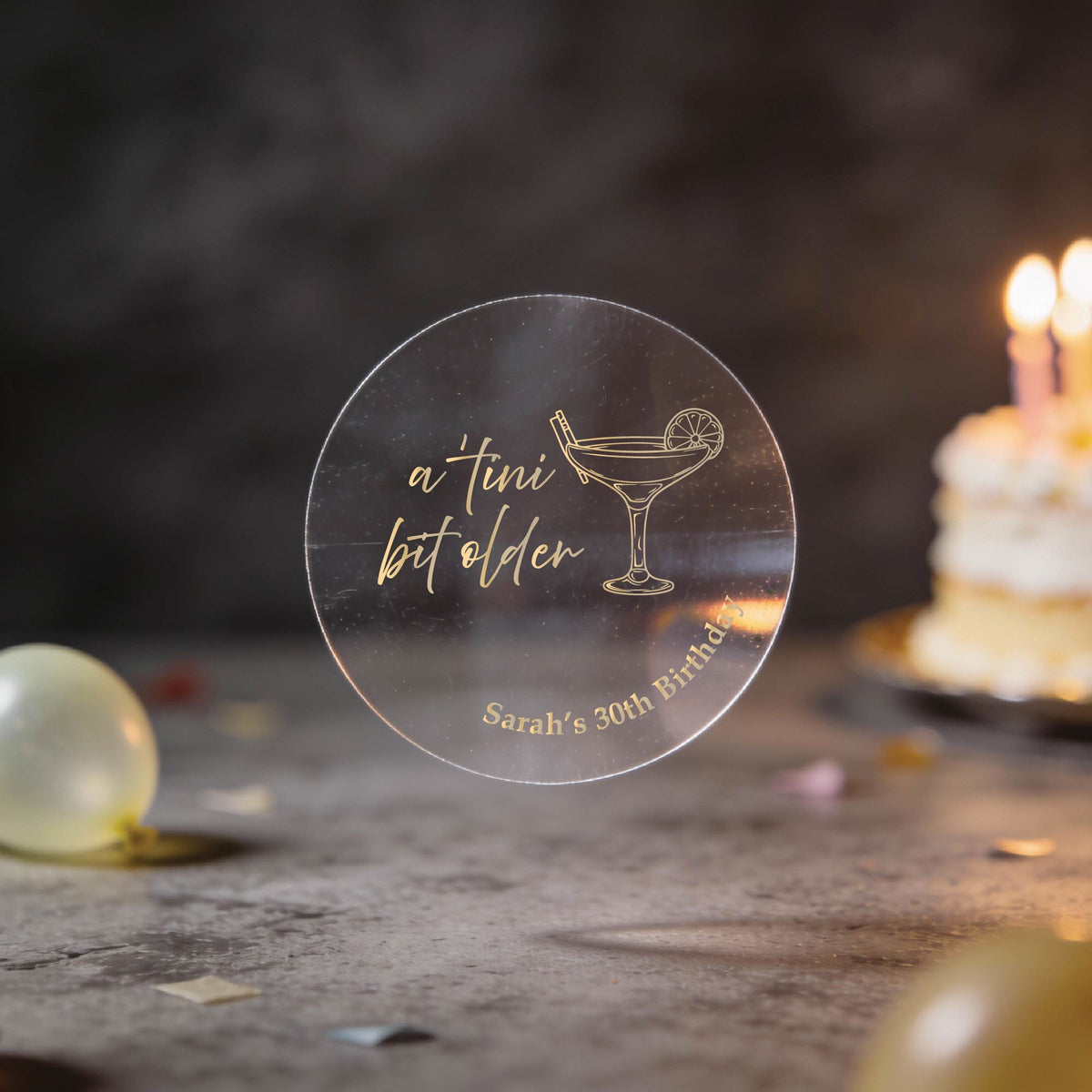 A round clear sticker with the words in gold foil saying 'a tini bit older' and a martini glass illustration, with 'Sarah's 30th birthday' printed at the bottom. The background is a dark party setting with blurred balloons and a birthday cake with lit candles.