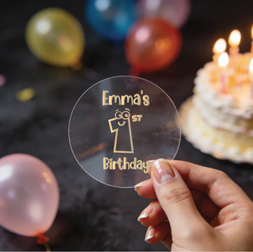 A hand holds a clear round gold foil sticker and the text 'Emma's 1st Birthday'. There is a large number 1 with eyes and a smile in the middle of the sticker.