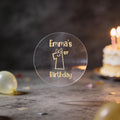 A clear round gold foil sticker and the text 'Emma's 1st Birthday'. There is a large number 1 with eyes and a smile in the middle of the sticker.