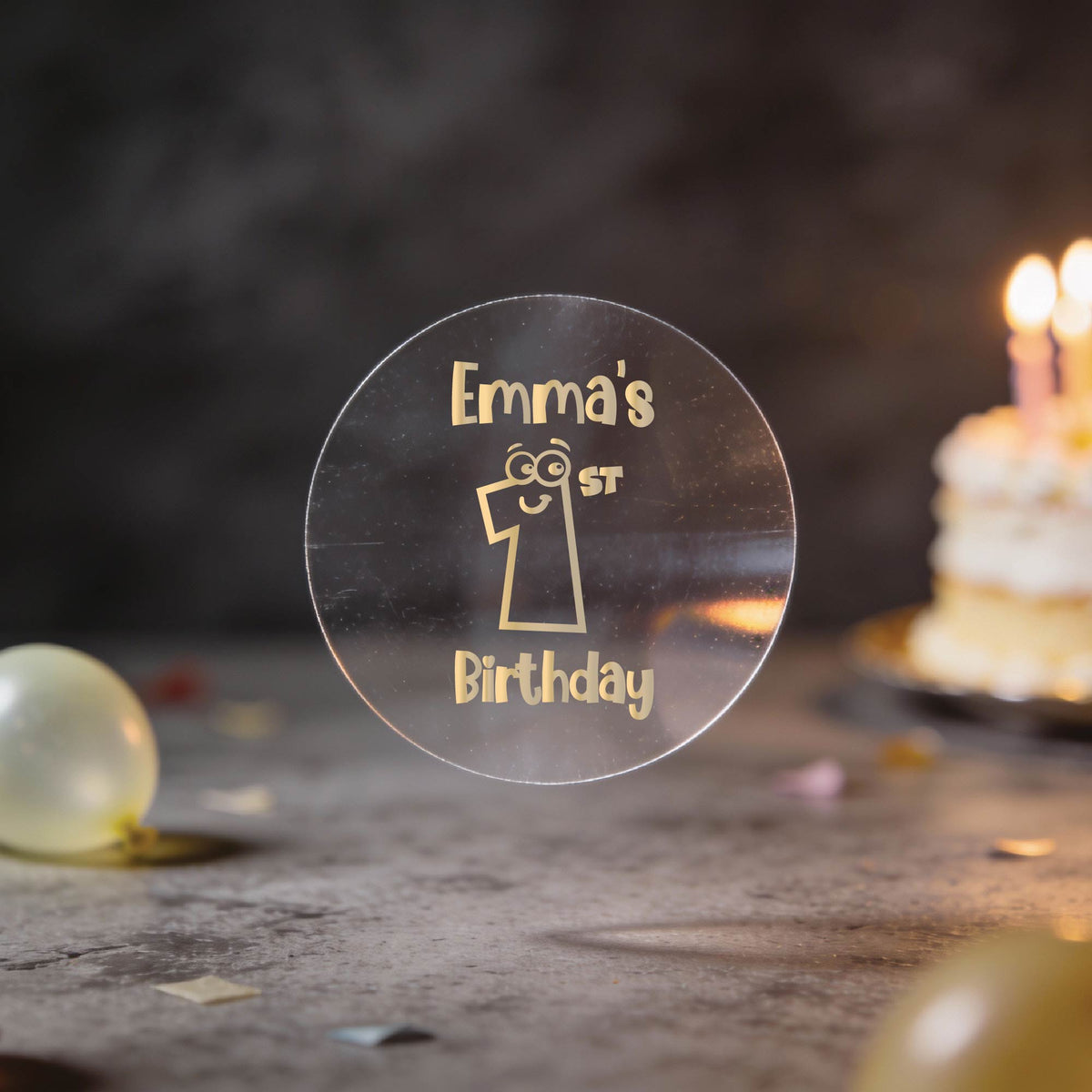 A clear round gold foil sticker and the text 'Emma's 1st Birthday'. There is a large number 1 with eyes and a smile in the middle of the sticker.