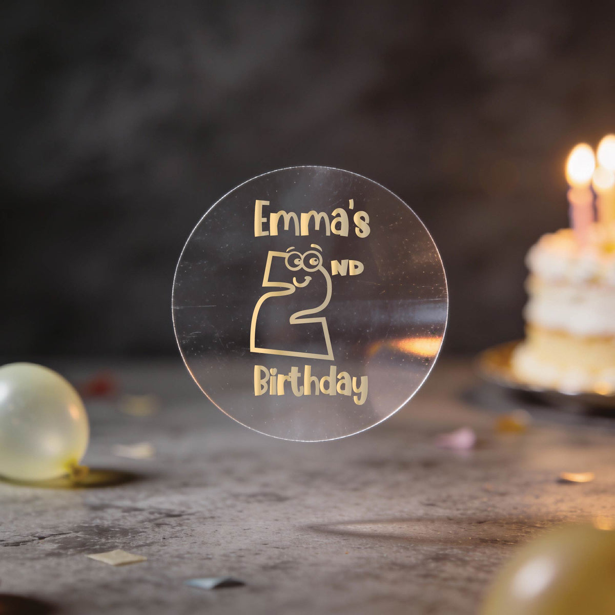 A clear round sticker with gold foil lettering that reads 'Emma's 2nd Birthday' and a cartoon number 2, shown on a tabletop with a blurred birthday cake and candles in the background.
