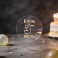 A clear round sticker with gold lettering that reads 'Emma's 3rd Birthday' stands on a dark tabletop. A blurred birthday cake with lit candles and confetti appear in the background, creating a celebratory setting.