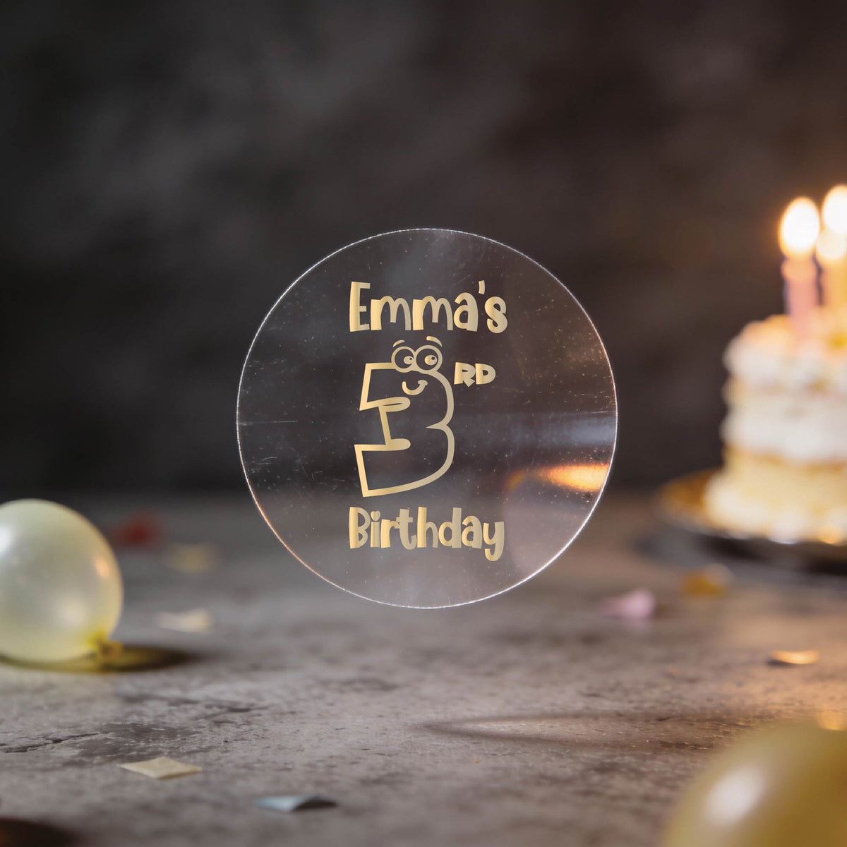 A clear round sticker with gold lettering that reads 'Emma's 3rd Birthday' stands on a dark tabletop. A blurred birthday cake with lit candles and confetti appear in the background, creating a celebratory setting.