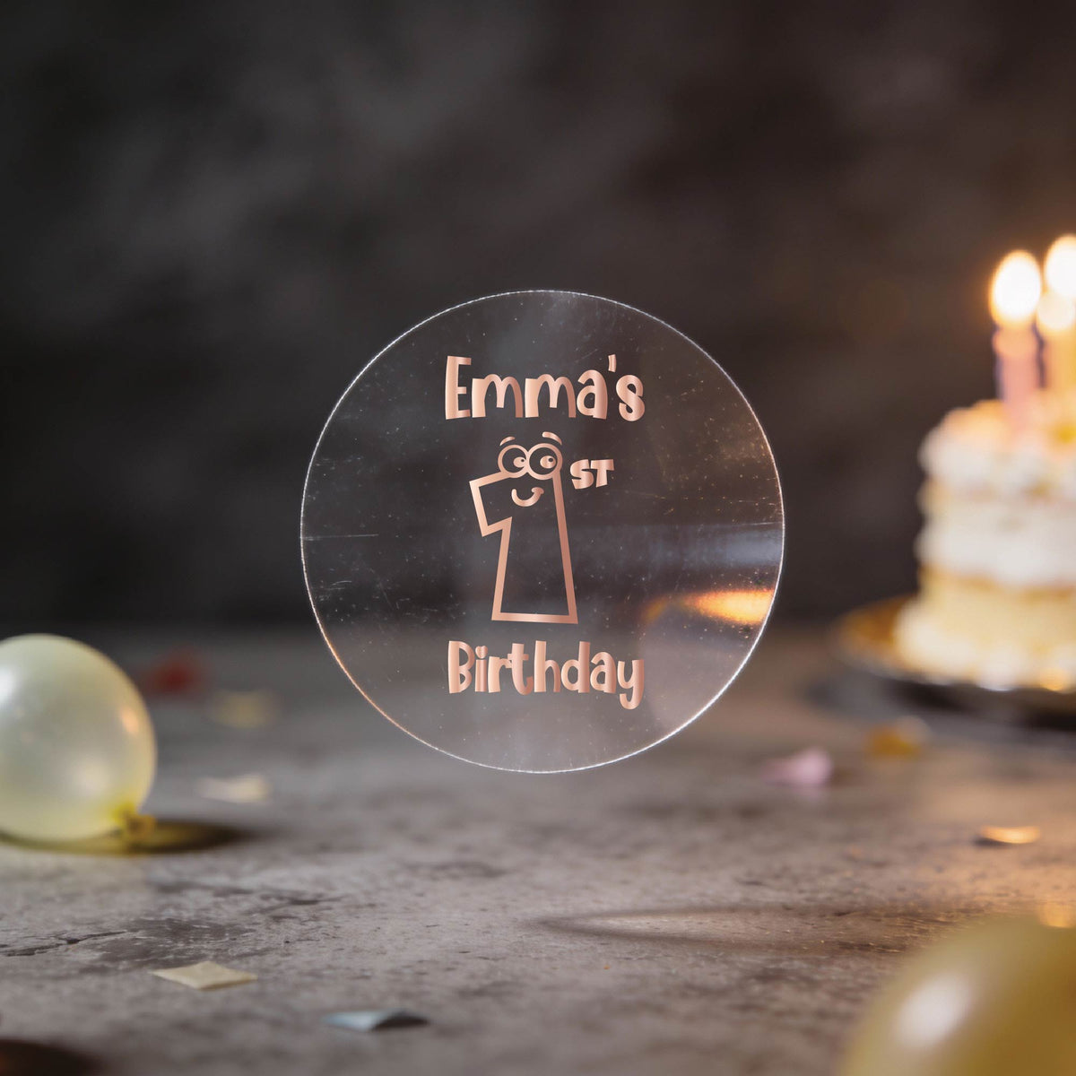A clear round rose gold foil sticker and the text 'Emma's 1st Birthday'. There is a large number 1 with eyes and a smile in the middle of the sticker.