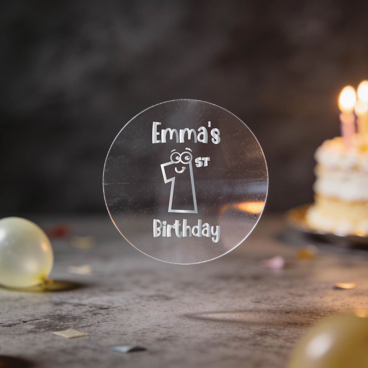 A clear round silver foil sticker and the text 'Emma's 1st Birthday'. There is a large number 1 with eyes and a smile in the middle of the sticker.