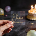 A hand holds a clear square sticker with gold foil text reading 'Olivia's 30th Birthday' against a dark birthday-themed background with blurred balloons and a lit cake.