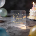 A clear square sticker with gold foil text reading 'Olivia's 30th Birthday' against a dark birthday-themed background with blurred balloons and a lit cake.