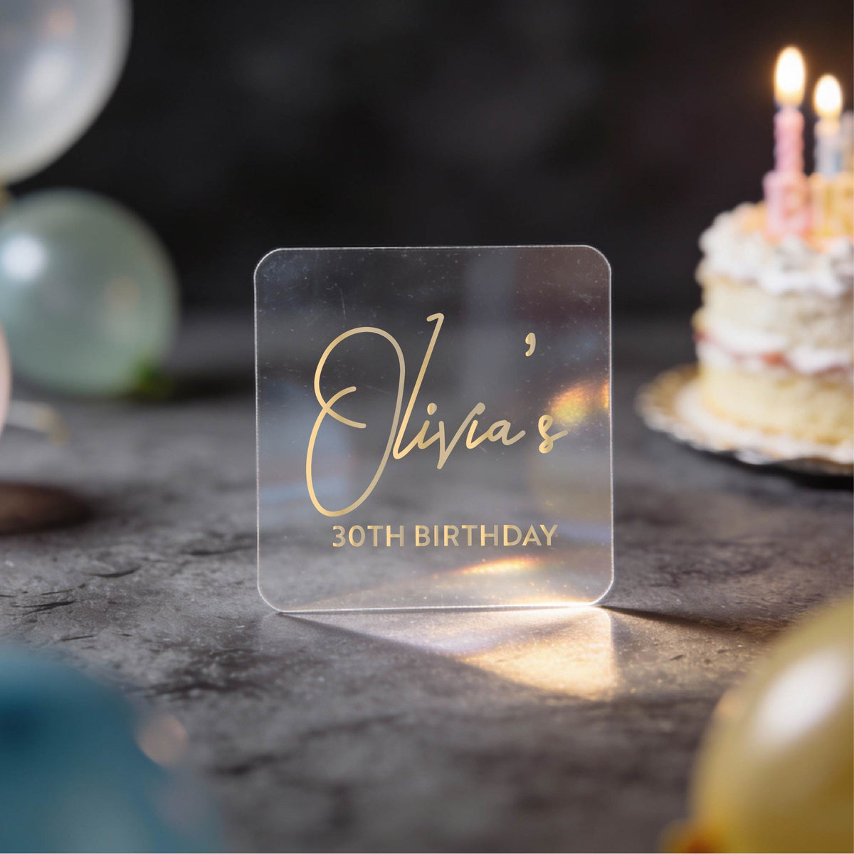 A clear square sticker with gold foil text reading 'Olivia's 30th Birthday' against a dark birthday-themed background with blurred balloons and a lit cake.