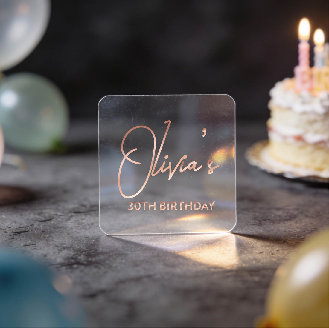 A hand holds a clear square sticker with gold foil text reading 'Olivia's 30th Birthday' against a dark birthday-themed background with blurred balloons and a lit cake.