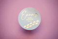 A round, white sticker with the text 'Baby Shower' and decorative elements, personalised with the name 'Emma' written in gold foil, on a pink background.