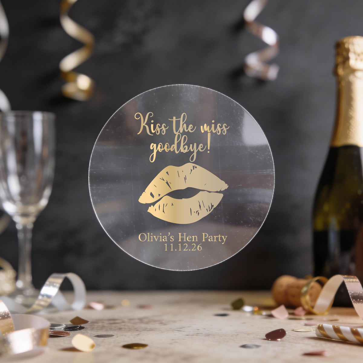A round clear sticker with gold foil text reading 'Kiss the miss goodbye!' and a gold lips graphic, displayed on a party table with a champagne bottle, glass, and confetti in the background.