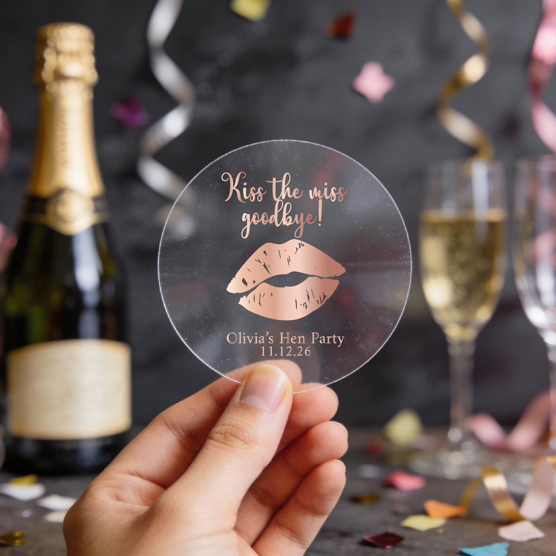 A round clear sticker with rose gold foil text reading 'Kiss the miss goodbye!' and a gold lips graphic, displayed on a party table with a champagne bottle, glass, and confetti in the background.
