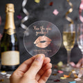 A round clear sticker with rose gold foil text reading 'Kiss the miss goodbye!' and a gold lips graphic, displayed on a party table with a champagne bottle, glass, and confetti in the background.