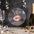 A round clear sticker with rose gold foil text reading 'Kiss the miss goodbye!' and a gold lips graphic, displayed on a party table with a champagne bottle, glass, and confetti in the background.