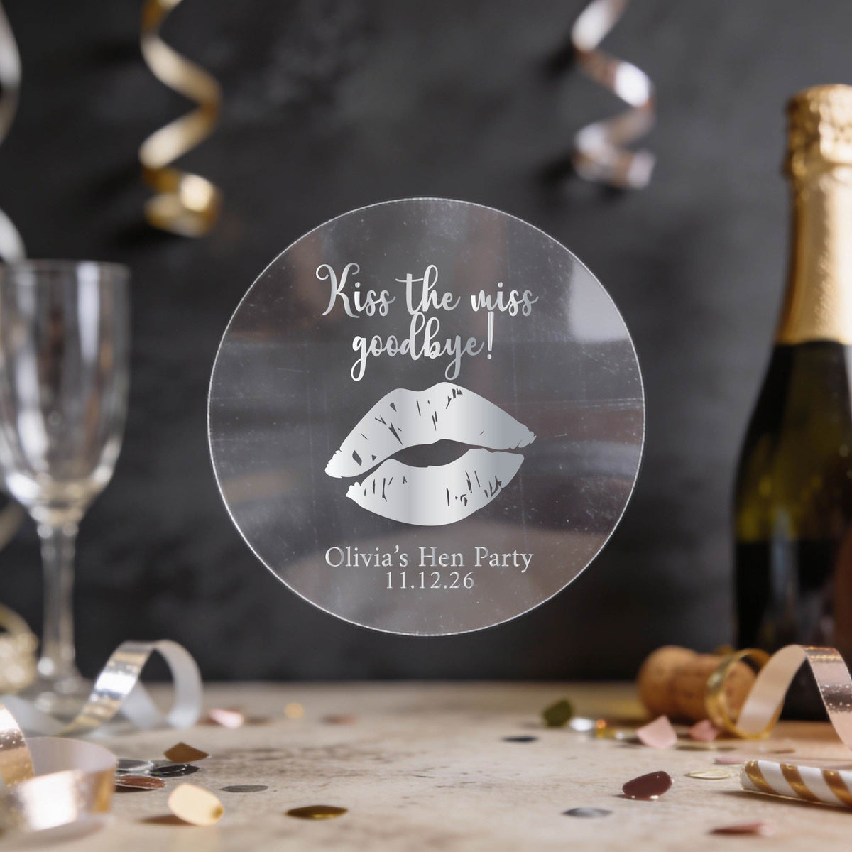 A round clear sticker with silver foil text reading 'Kiss the miss goodbye!' and a gold lips graphic, displayed on a party table with a champagne bottle, glass, and confetti in the background.