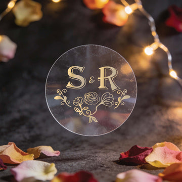A round, clear sticker with gold foil initials "S & R" and floral design in the centre. Surrounded by fairy lights and scattered rose petals on a dark surface.
