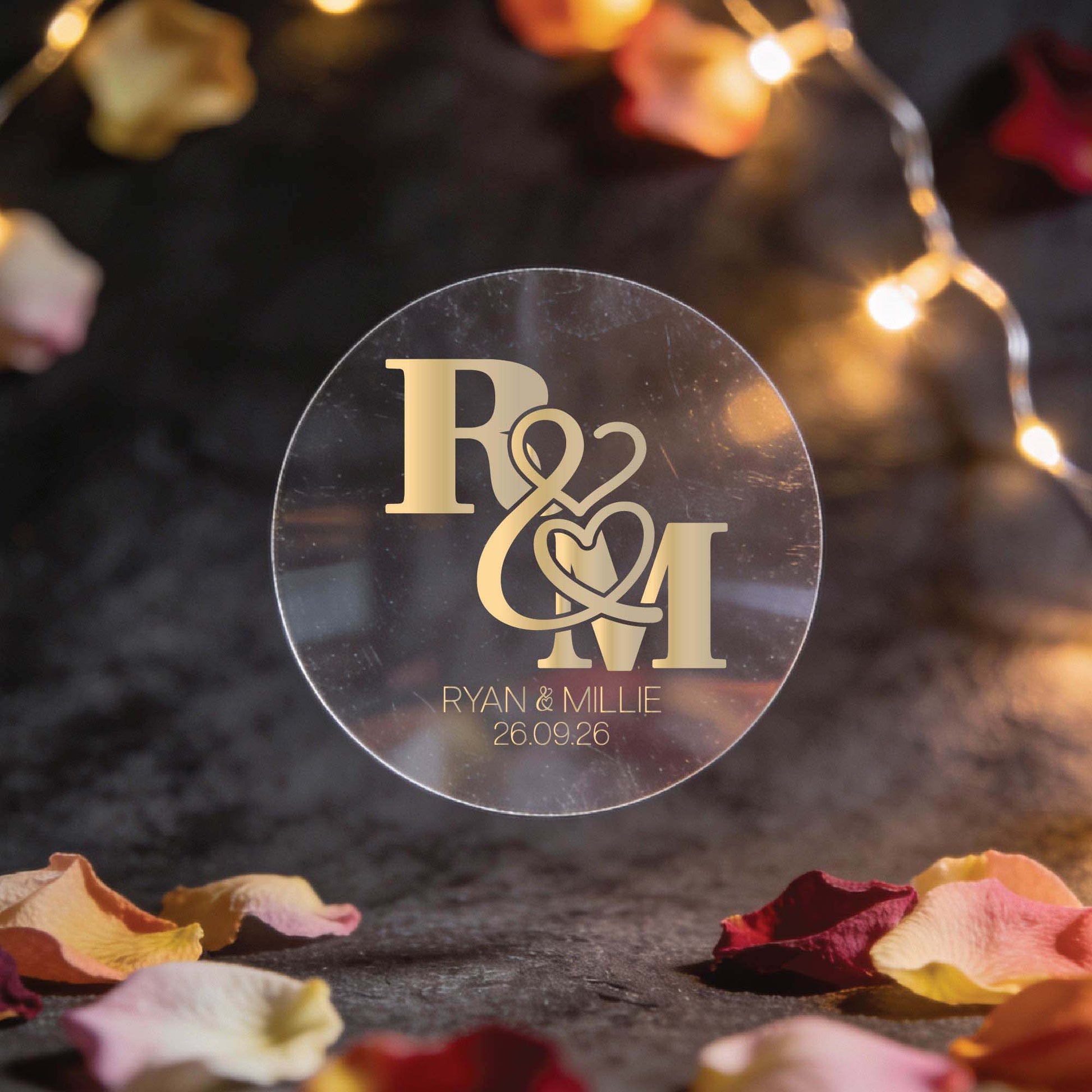 A circular transparent sticker with gold foil initials "R & M" intertwined with heart motifs. Below, "Ryan & Millie, 26.09.26." Surrounded by fairy lights and rose petals, creating a romantic ambiance.