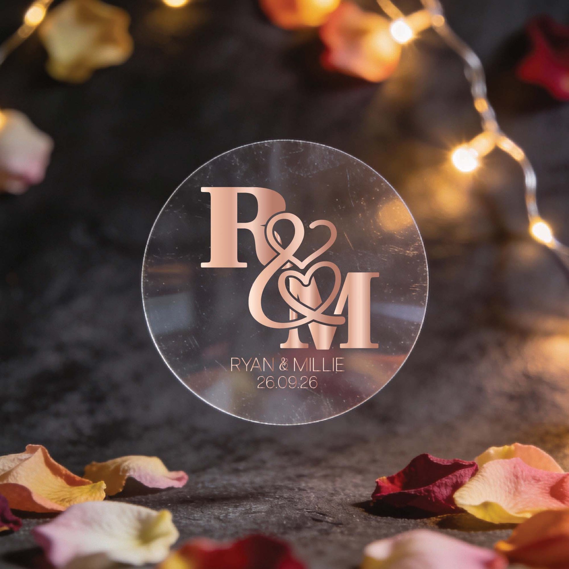 A circular transparent sticker with rose gold foil initials "R & M" intertwined with heart motifs. Below, "Ryan & Millie, 26.09.26." Surrounded by fairy lights and rose petals, creating a romantic ambiance.