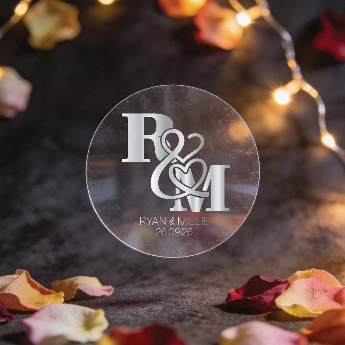 A circular transparent sticker with gold foil initials "R & M" intertwined with heart motifs. Below, "Ryan & Millie, 26.09.26." Surrounded by fairy lights and rose petals, creating a romantic ambiance.