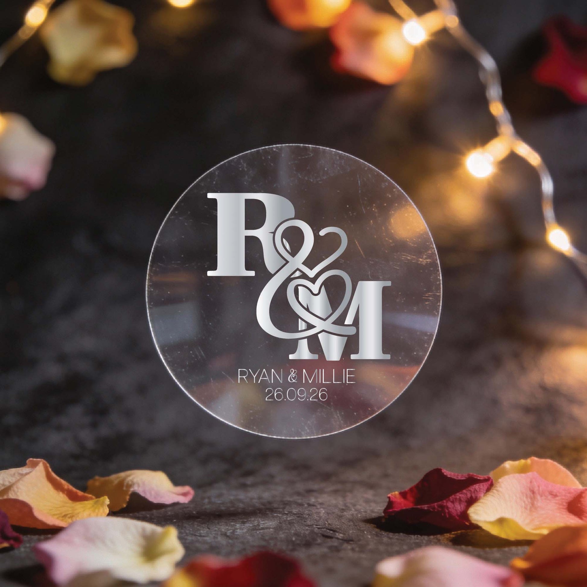 A circular transparent sticker with silver foil initials "R & M" intertwined with heart motifs. Below, "Ryan & Millie, 26.09.26." Surrounded by fairy lights and rose petals, creating a romantic ambiance.